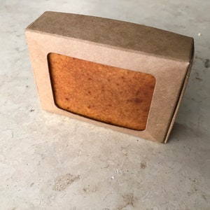 May include: A rectangular bar of soap in a brown cardboard box with a rectangular window. The soap is a light brown colour with a slightly textured surface. The box is made of recycled cardboard.