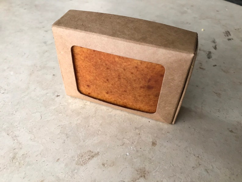 May include: A rectangular bar of soap in a brown cardboard box with a rectangular window. The soap is a light brown colour with a slightly textured surface. The box is made of recycled cardboard.