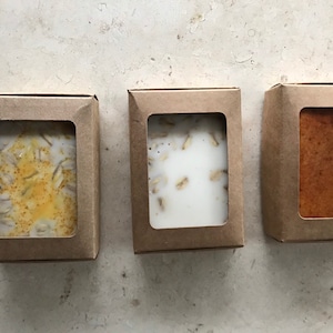May include: Three bars of soap in cardboard boxes with clear windows. The soap bars have different textures and colors, including a light brown bar, a bar with oats, and a bar with yellow specks.