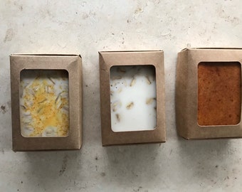 May include: Three soap bars in cardboard boxes with clear windows. The soap bars have different textures and colours, including a light brown bar, a bar with oats, and a bar with yellow specks.