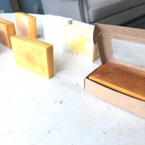 May include: A selection of rectangular handmade soap bars in shades of yellow and brown are displayed on a white surface. One bar is presented in an open cardboard box with a clear window. The soaps have a natural, rustic appearance.