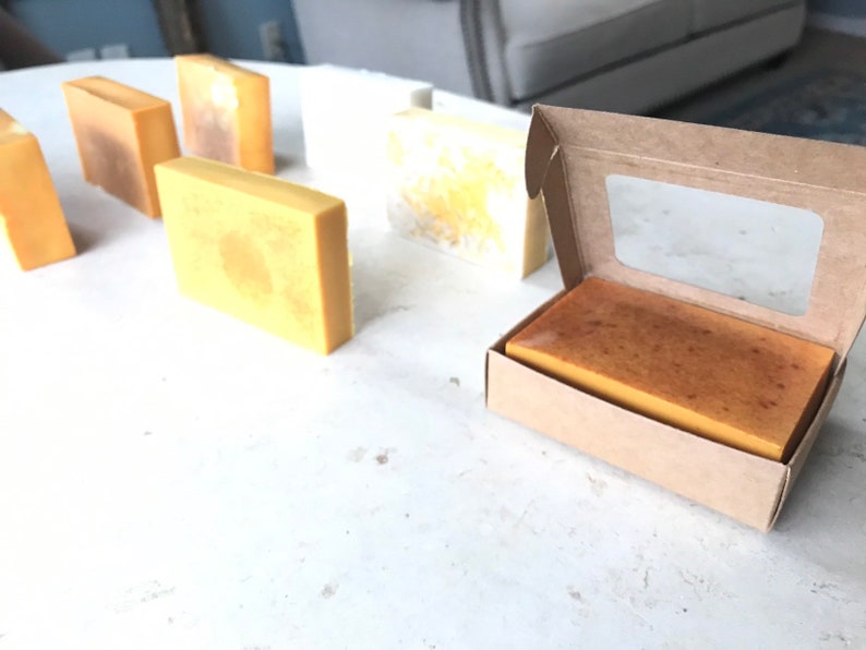 May include: A selection of rectangular handmade soap bars in shades of yellow and brown are displayed on a white surface. One bar is presented in an open cardboard box with a clear window. The soaps have a natural, rustic appearance.