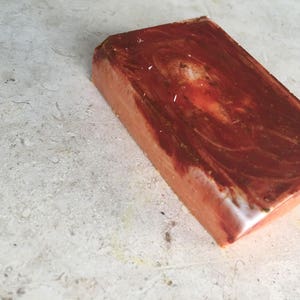 May include: A rectangular bar of handmade soap with a marbled, reddish-brown surface. The soap has a light orange base colour with darker brown swirls and a white edge. The soap is on a light grey surface.