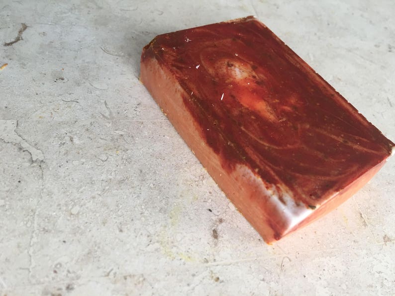 May include: A rectangular bar of handmade soap with a marbled, reddish-brown surface. The soap has a light orange base colour with darker brown swirls and a white edge. The soap is on a light grey surface.
