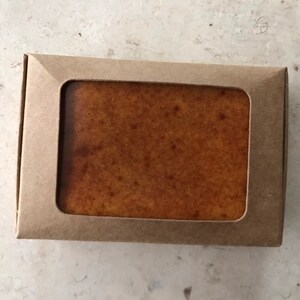 May include: A rectangular bar of soap in a brown cardboard box with a rectangular window. The soap is a light brown colour with darker brown speckles. The box is a light brown colour.