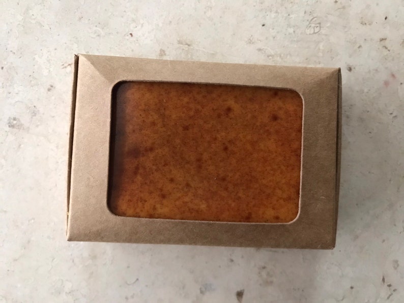 May include: A rectangular bar of soap in a brown cardboard box with a rectangular window. The soap is a light brown colour with darker brown speckles. The box is a light brown colour.
