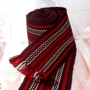 May include: A red, white, and brown woven fabric strap with fringe on the ends. The strap is rolled up and tied in a knot.