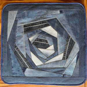 May include: A square cushion cover constructed from various shades of blue denim fabric strips arranged in a spiraling, geometric pattern. The edges are finished with a dark blue piped border, creating a textured, layered appearance throughout.