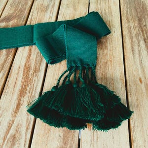 Pode incluir: Uma faixa verde escura com uma longa franja de borlas. A faixa é feita de um tecido tecido e tem uma textura lisa.