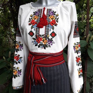 May include: A white linen blouse with embroidered floral and geometric patterns in red, yellow, blue, and black. The blouse has a red tassel and is worn with a red and black woven belt.