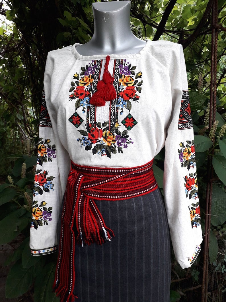 May include: A white linen blouse with embroidered floral and geometric patterns in red, yellow, blue, and black. The blouse has a red tassel and is worn with a red and black woven belt.