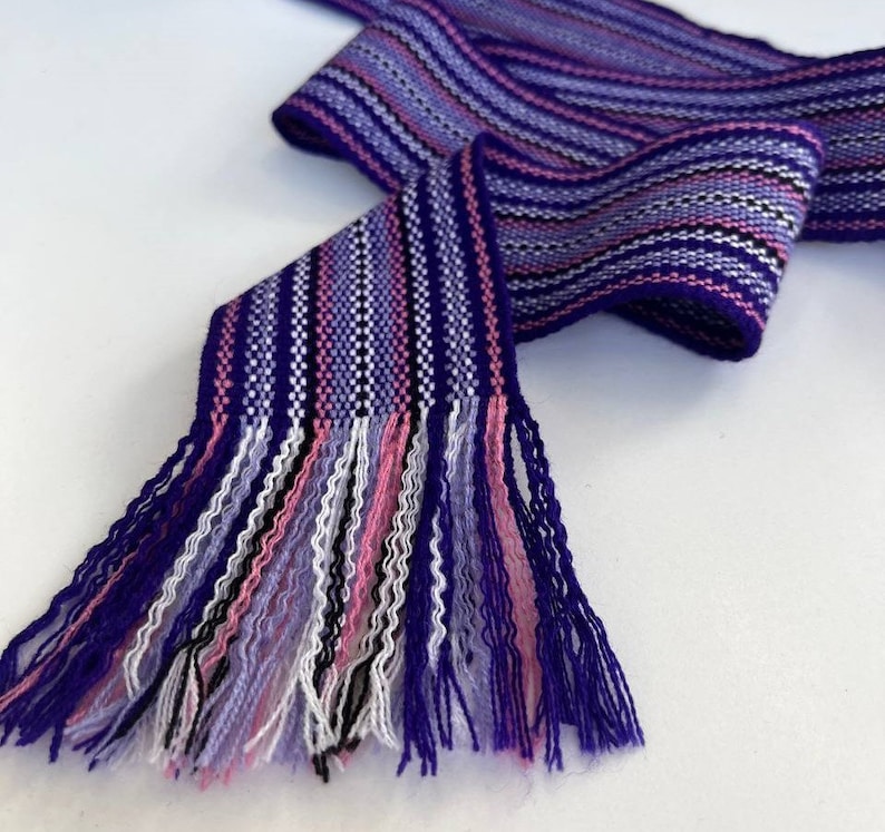 May include: A purple, pink, white, and black striped woven fabric with fringe on one end. The fabric is folded in half and the fringe is hanging down.