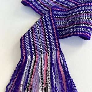May include: A purple, pink, white, and black woven fabric ribbon with fringe tassels. The ribbon is striped and has a woven pattern.