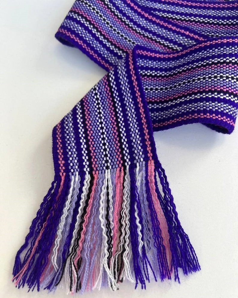 May include: A purple, pink, white, and black woven fabric ribbon with fringe tassels. The ribbon is striped and has a woven pattern.