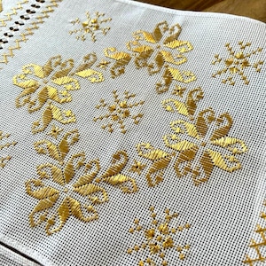 May include: Close-up of a white embroidered table runner with a repeating pattern of gold floral and snowflake designs. The edges feature a decorative border with a cut-out design and gold stitching.