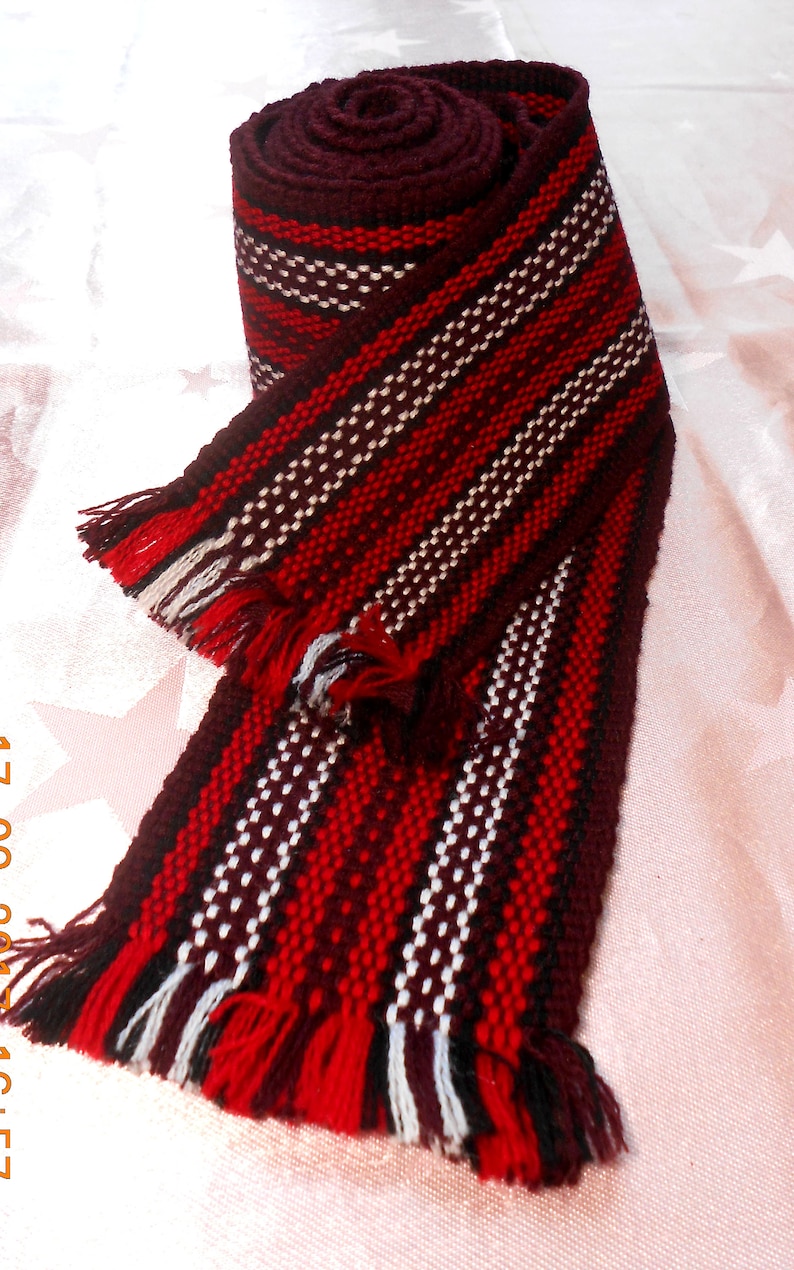 May include: A red, white, and brown woven fabric strap with fringe on the ends. The strap is rolled up and tied in a knot.