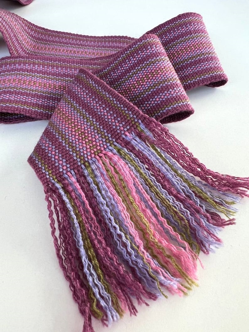 May include: A woven purple, pink, and green fabric with a fringe. The fabric has a geometric pattern.