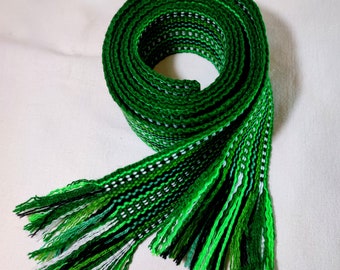 May include: A green, white, and black woven belt with fringe on the ends. The belt is coiled into a circle.