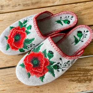 以下が含まれることがあります： 赤いポピーと緑の葉が刺繍された、明るい灰色のフェルトスリッパのペア。 スリッパの縁には赤いステッチが施されています。 このスリッパは屋内での使用を目的としています。