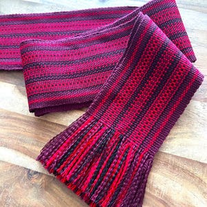 Pode incluir: Um cachecol tecido à mão em tons de vermelho e preto. O cachecol apresenta um padrão listrado com uma borda com franjas. As cores são profundas e ricas, criando um acessório acolhedor. O cachecol é feito de um material macio e texturizado.