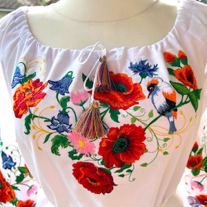 Könnte beinhalten: Weiß bestickte Bluse mit einem lebendigen Blumenmuster mit roten Mohnblumen, blauen Kornblumen und einem bunten Vogel. Die Bluse hat einen runden Ausschnitt und Quasten Details, im Folk-Art-Stil.