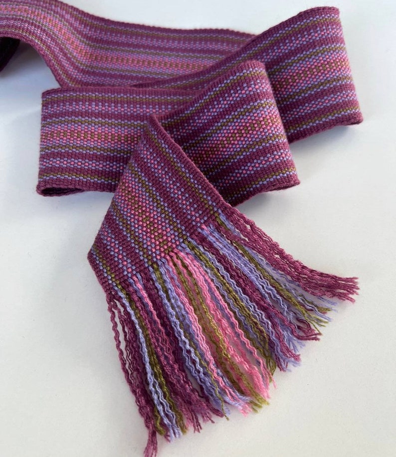 May include: A woven purple, pink, and green striped ribbon with a fringe on one end.