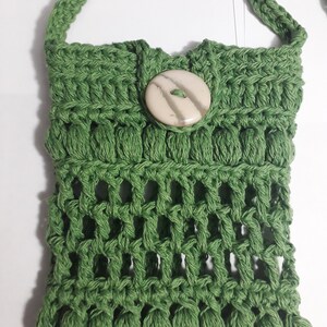 Puede incluir: Un bolso de hombro de crochet verde con cierre de botón blanco. El bolso tiene un diseño tejido y una correa larga.