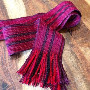 Pode incluir: Cachecol tecido à mão em tons de vermelho e preto, com um padrão listrado. O cachecol tem uma borda com franjas, adicionando textura. O tecido é exibido em uma superfície de madeira.