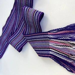 May include: A purple, pink, white, and black woven fabric ribbon with fringe on one end.