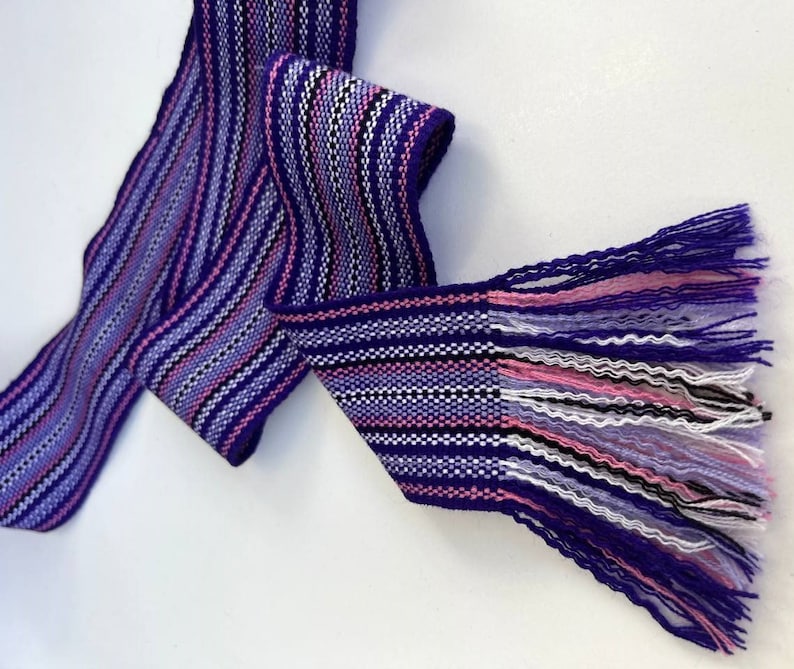 May include: A purple, pink, white, and black woven fabric ribbon with fringe on one end.