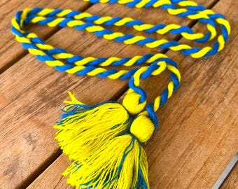 May include: A decorative tassel and cord set in yellow and blue. The cord is twisted, and the tassel features a large, fluffy design. The colors are vibrant against the wooden surface.