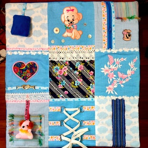 Puede incluir: Una manta de actividad para bebés multicolor con varias texturas y patrones. La manta presenta un corazón, un perro, una flor, una cremallera y un ribete de encaje.