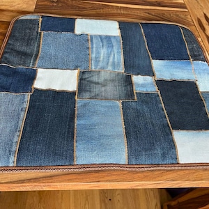 May include: A rectangular denim patchwork cushion featuring various shades of blue denim squares. The cushion has a brown border and is placed on a wooden surface. The design showcases a mix of light and dark denim pieces.