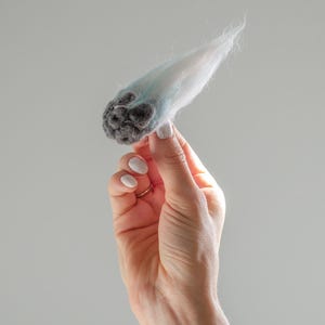 May include: A handmade felted sculpture in shades of gray, white, and light blue. The sculpture has a rounded, textured base and a wispy, elongated top. The object is held in a hand against a plain gray background.