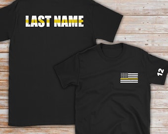 May include: Two black t-shirts with a thin yellow stripe. The back of one shirt has the text "LAST NAME" in white. The front of the other shirt has a small American flag with a thin yellow stripe across it. The sleeve of the second shirt has the number "12" in white.