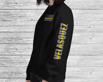 May include: Black sweatshirt with a thin yellow stripe down the right sleeve. The stripe has the word "VELLASQUEZ" written in gray letters. A small American flag with a thin yellow stripe is on the left chest.