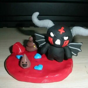 Pode incluir: Uma pequena figura de argila de uma criatura preta com chifres e asas cinzentas, olhos brancos e marcas vermelhas. Fica sobre uma base circular vermelha ao lado de uma pequena figura cónica castanha sorridente, uma rocha vermelha e três pequenas peças azuis em forma de coração.