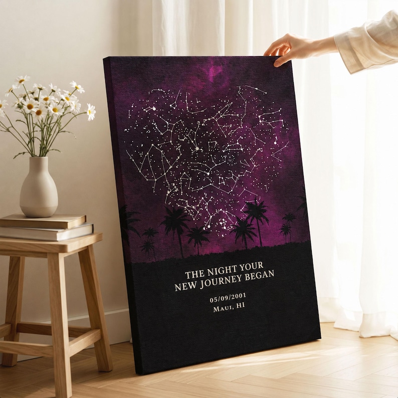 A person's hand is holding up a canvas print of a starry night sky with palm trees silhouetted against it, placed on a wooden table next to a vase of white flowers.