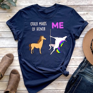 May include: A navy blue t-shirt with the text "Other maids of honour" and "Me" printed on it. The text "Me" is in pink and is next to a unicorn doing a pole dance. The text "Other maids of honour" is next to a brown horse.