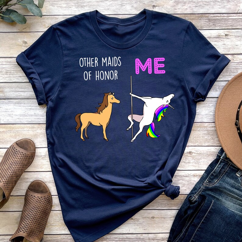 May include: A navy blue t-shirt with the text "Other maids of honour" and "Me" printed on it. The text "Me" is in pink and is next to a unicorn doing a pole dance. The text "Other maids of honour" is next to a brown horse.