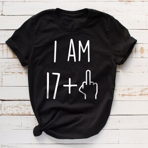 May include: Black t-shirt with white text that reads "I AM 17+" with a middle finger emoji.