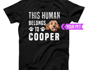 May include: Black t-shirt with the text "THIS HUMAN BELONGS TO COOPER" in white. The design includes paw prints and a photo of a brown dog. A purple banner on the right says "YOUR PET".