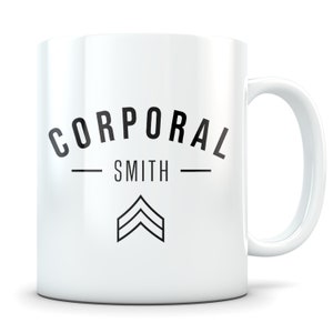 May include: White ceramic coffee mug with black text that reads "Corporal Smith" with a black chevron symbol below the text.