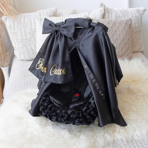 Peut inclure: Canopée de siège auto en satin noir avec un nom brodé en or "Olivia Cava" et une doublure en fausse fourrure noire. La canopée a un nœud en haut.