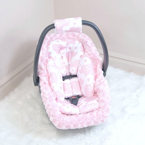 May include: Pink baby car seat cover with a white and pink floral pattern. The cover is made of soft, plush fabric and has a headrest and a strap for added comfort.