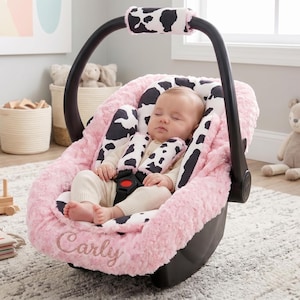 May include: A baby car seat with a pink, plush exterior and cow print accents. The seat features a black handle and a sleeping baby secured with a harness. The name "Carly" is embroidered on the seat.