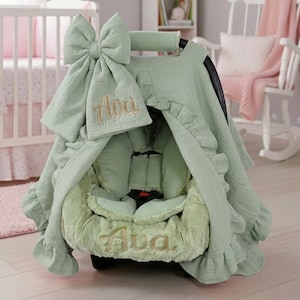 May include: A sage green baby car seat cover with a matching bow and hat. The name "Ava" is embroidered in gold thread on the hat and seat cover. The cover has a ruffled edge and a plush interior.