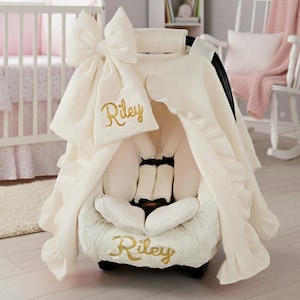 May include: Ivory-colored baby car seat cover with a large bow and ruffle trim. The name "Riley" is embroidered in gold script on the cover and matching headrest. The seat is designed for infant use and includes safety straps.