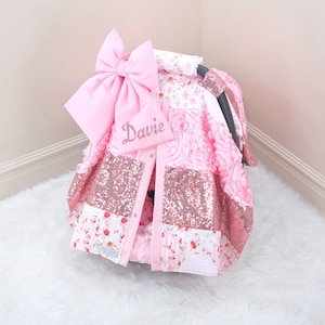 May include: Pink and rose gold car seat cover with a large pink bow and the name "Davie" embroidered on it.