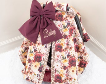 May include: A burgundy and gold car seat cover with a floral pattern and a large burgundy bow. The cover is personalized with the name "Bailey".